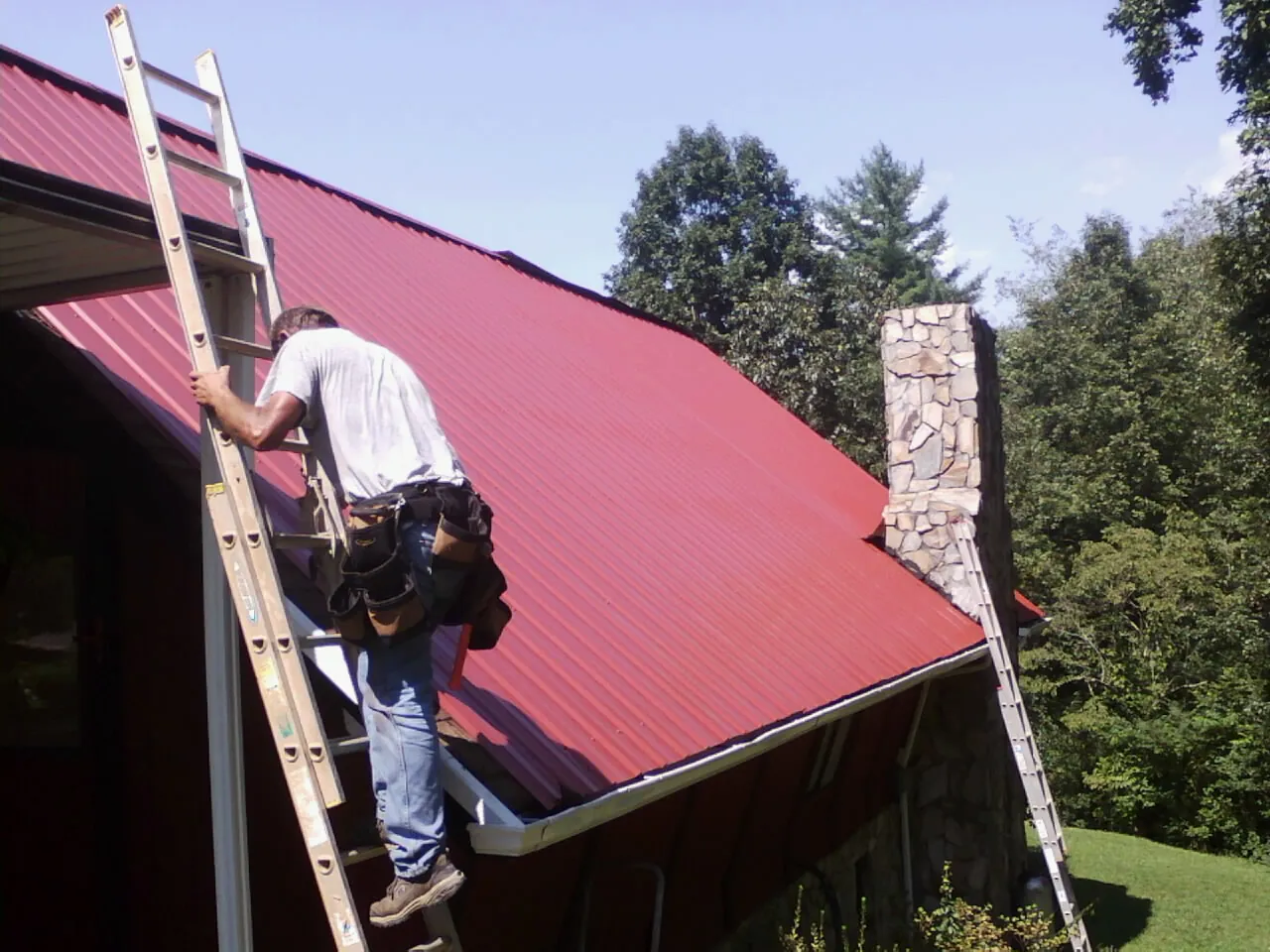 Roofer on a ladder beside a red metal roof and stone chimney — Soffit & Fascia Repair by Roofing Brothers in Manchester Township